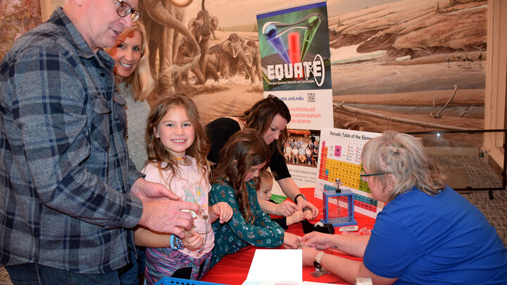 Girl smiles at camera while family is engaged in STEM activity. 